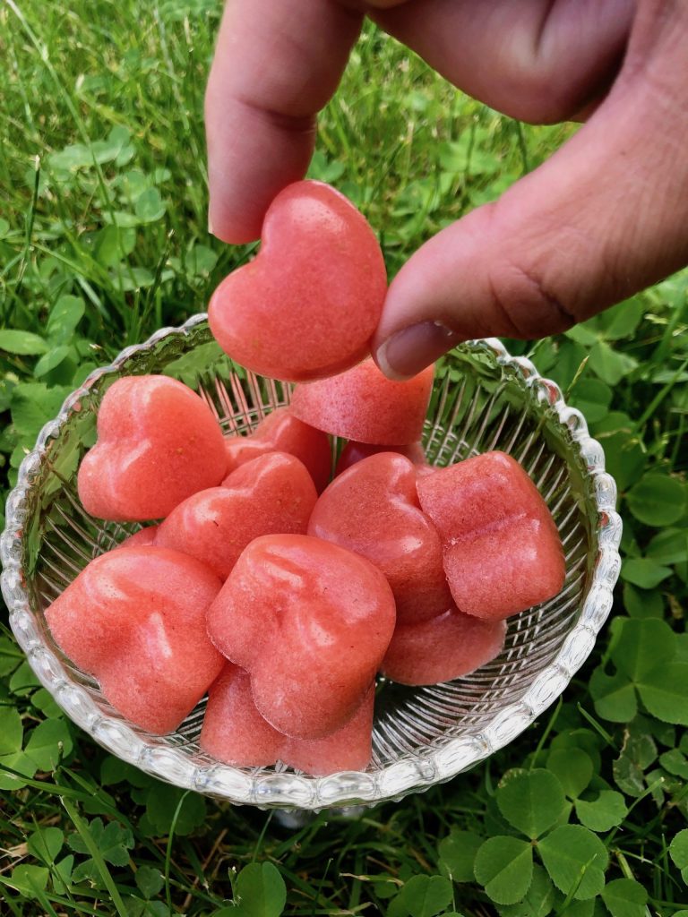 Bild på någon som plockar upp en gelégodis, gjord på jordgubbar, från en skål. Gelegodisbitarna är härligt röda och hjärtformade. Bilden är tagen utomhus på en gräsmatta.