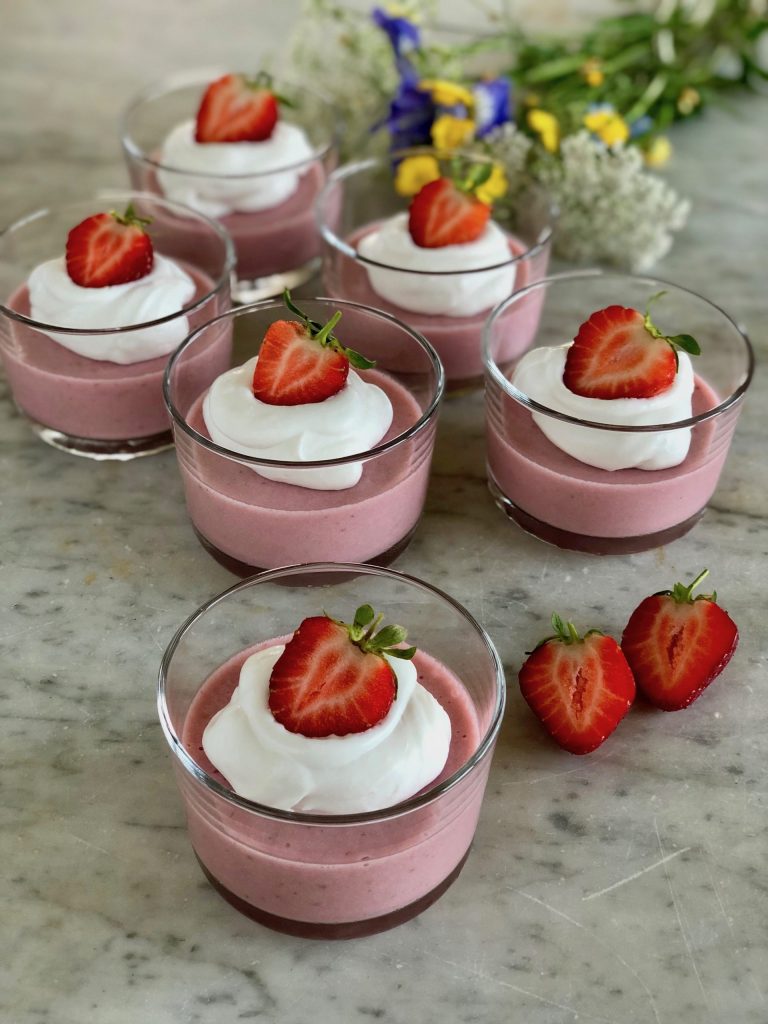 Bild på jordgubbsmousse med kokosgrädde där en halverad jordgubbe toppar desserten. Det är totalt 6 glas med mousse och i bakgrunden ligger en bucket med midsommarblomster.